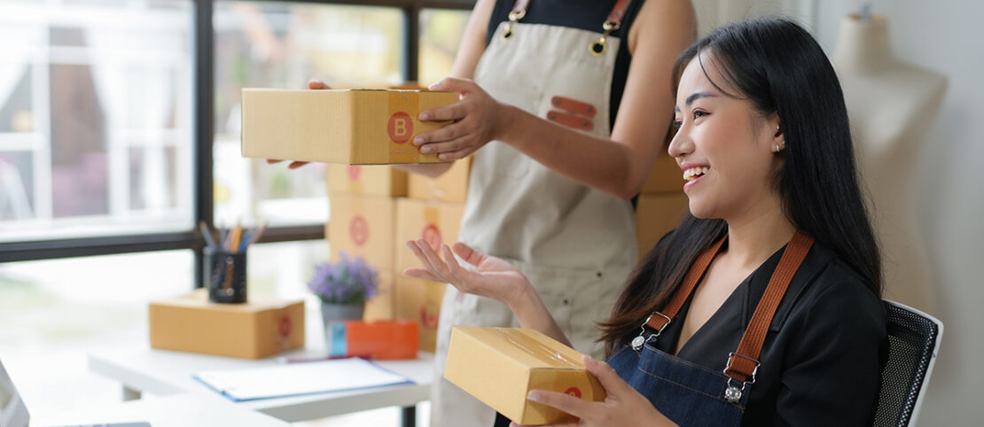 Two young women are collaborating, managing their online business from their home office. They are smiling while packing orders